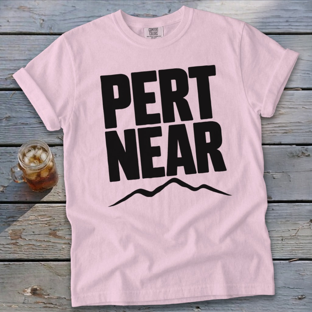 Light pink t-shirt featuring the black text PERT NEAR above a mountain range graphic, resting on a weathered wooden surface next to an iced beverage.