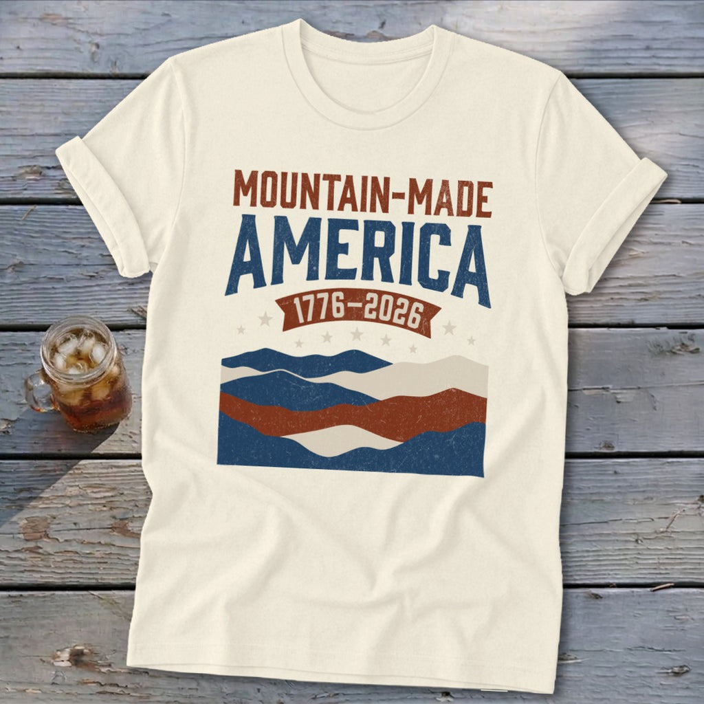 Cream t-shirt featuring a distressed red, white, and blue mountain graphic with the text Mountain-Made America 1776-2026.