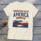 Cream t-shirt featuring a distressed red, white, and blue mountain graphic with the text Mountain-Made America 1776-2026.