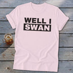 A light pink crewneck t-shirt with rolled sleeves lies flat on a gray wooden surface next to a glass of iced tea. The shirt features the black text "WELL I SWAN" centered on the chest.