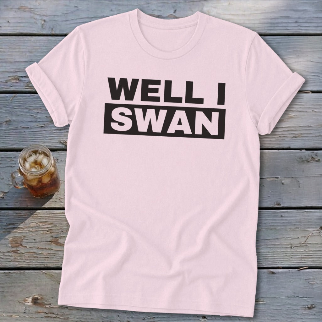 A light pink crewneck t-shirt with rolled sleeves lies flat on a gray wooden surface next to a glass of iced tea. The shirt features the black text "WELL I SWAN" centered on the chest.