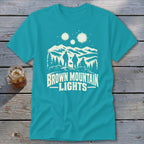 Turquoise t-shirt featuring a white graphic of three figures, mountains, glowing orbs, and text reading BROWN MOUNTAIN LIGHTS.