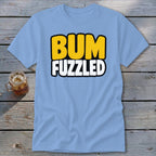 Light blue t-shirt featuring the text "BUM FUZZLED" in yellow and white, lying on a wooden surface beside a glass of iced tea.
