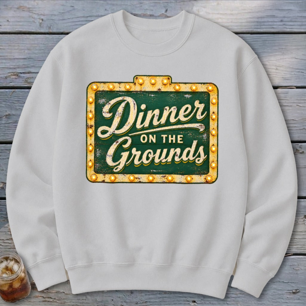 A white crewneck sweatshirt featuring a graphic of a vintage marquee sign reading Dinner on the Grounds.