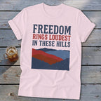 A light pink t-shirt with rolled sleeves features a graphic of red, blue, and dark blue mountains and the text "FREEDOM RINGS LOUDEST IN THESE HILLS."