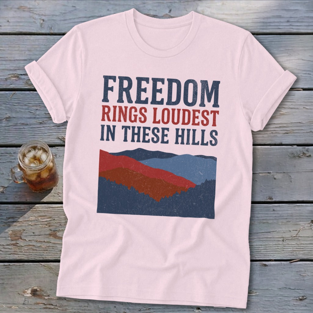 A light pink t-shirt with rolled sleeves features a graphic of red, blue, and dark blue mountains and the text "FREEDOM RINGS LOUDEST IN THESE HILLS."