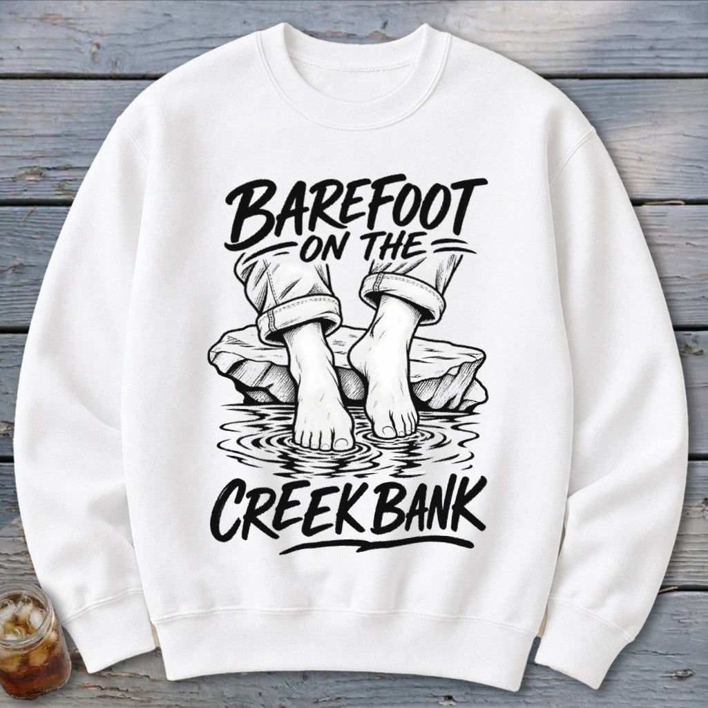White sweatshirt with a black graphic of bare feet on a rock in water, text reads "Barefoot on the Creek Bank."