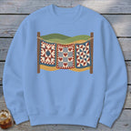 Light blue crewneck sweatshirt featuring a graphic of three patchwork quilts hanging on a clothesline against a landscape backdrop.