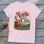A light pink t-shirt featuring a graphic of an apple, a cabin, and mountains with the text Apple At Cha.