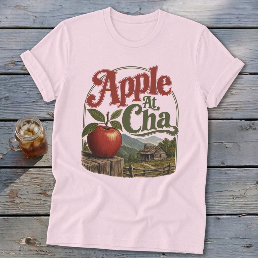 A light pink t-shirt featuring a graphic of an apple, a cabin, and mountains with the text Apple At Cha.