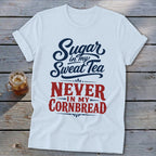 A white crewneck t-shirt with rolled sleeves featuring blue and red graphic text that reads Sugar in my Sweat Tea NEVER IN MY CORNBREAD laid flat on a rustic wooden background next to a glass of iced tea.