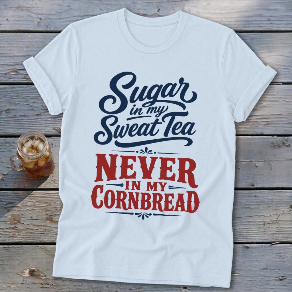 A white crewneck t-shirt with rolled sleeves featuring blue and red graphic text that reads Sugar in my Sweat Tea NEVER IN MY CORNBREAD laid flat on a rustic wooden background next to a glass of iced tea.
