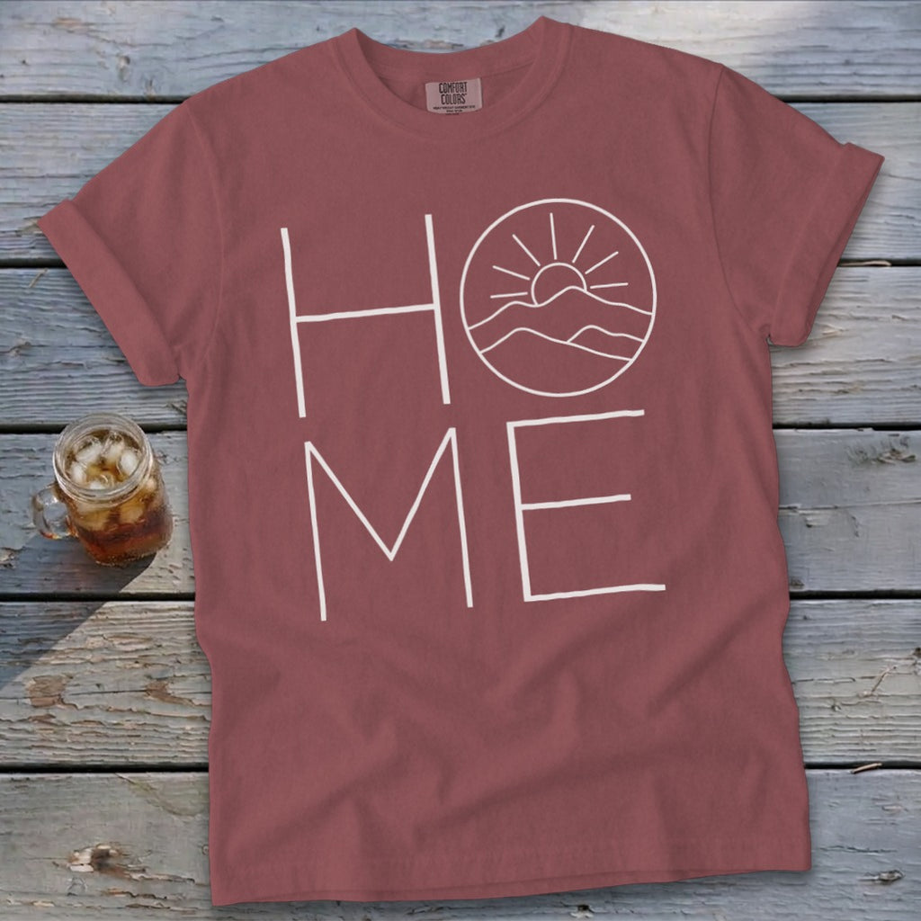 A brick red t-shirt with white text reading HOME, where the O is replaced by a sun and mountains illustration.