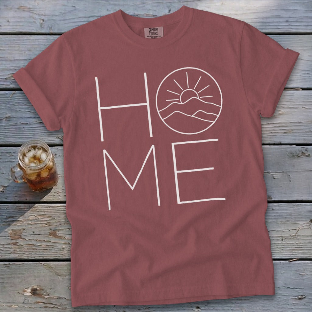 A brick red t-shirt with white text reading HOME, where the O is replaced by a sun and mountains illustration.