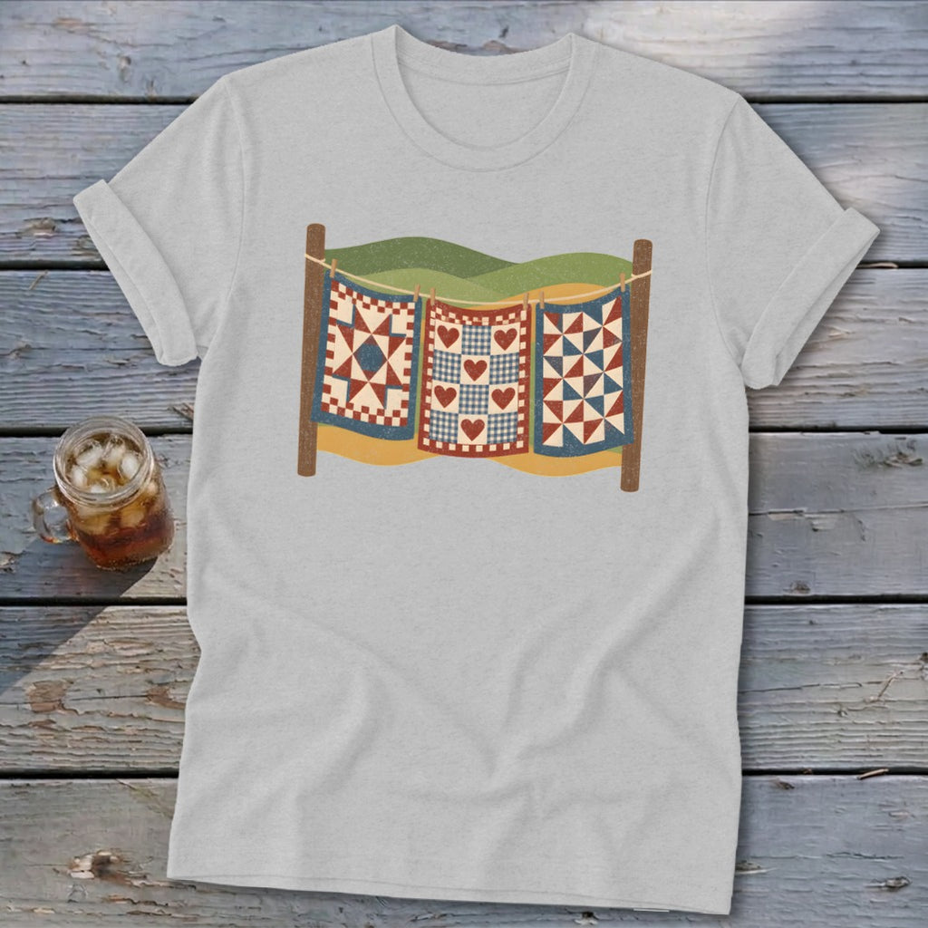 A light gray short-sleeve t-shirt features a vintage-style graphic of three colorful quilts hanging on a clothesline.