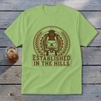 Light green t-shirt with a circular emblem featuring a lantern and the text "ESTABLISHED IN THE HILLS".