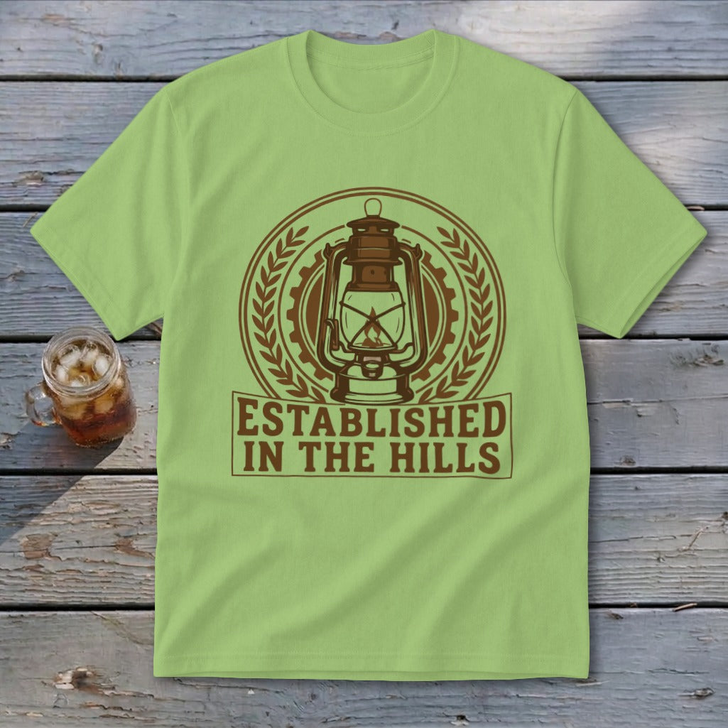 Light green t-shirt with a circular emblem featuring a lantern and the text "ESTABLISHED IN THE HILLS".