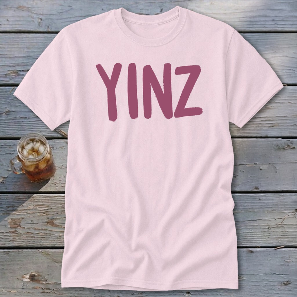 Pink t-shirt with maroon "YINZ" text on wooden surface next to glass of iced tea.
