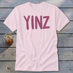 Pink t-shirt with maroon "YINZ" text on wooden surface next to glass of iced tea.
