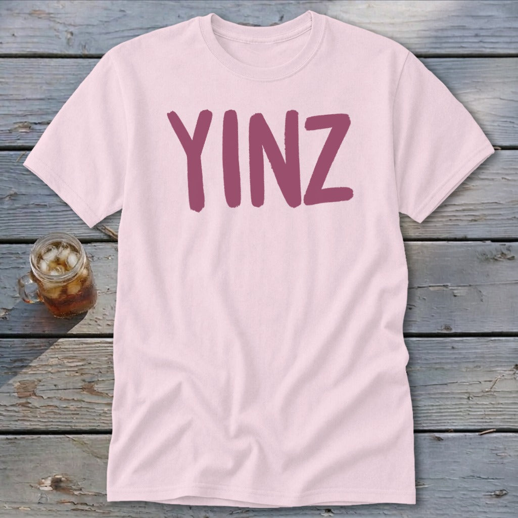 Pink t-shirt with maroon "YINZ" text on wooden surface next to glass of iced tea.