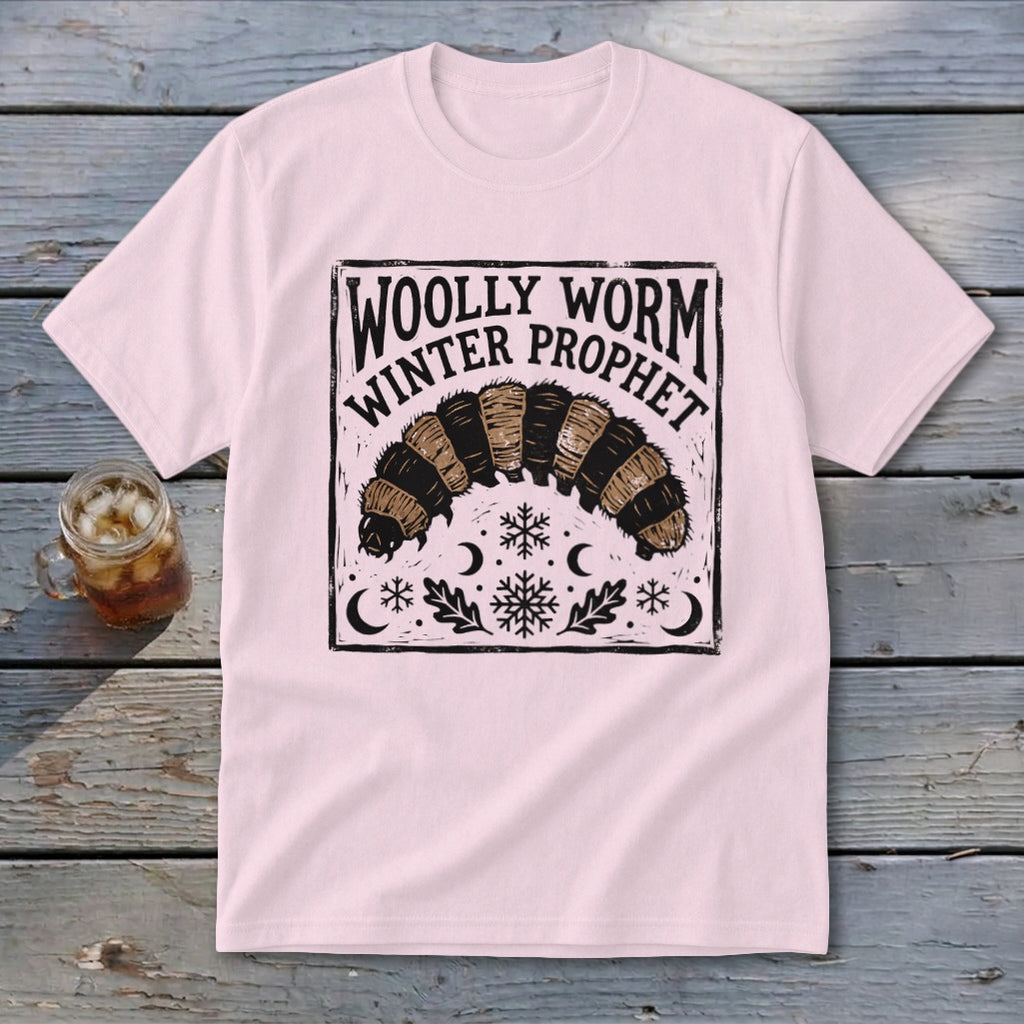 A light pink t-shirt featuring a black block-print design of a woolly worm and snowflakes with the text WOLLY WORM WINTER PROPHET.