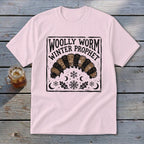 A light pink t-shirt featuring a black block-print design of a woolly worm and snowflakes with the text WOLLY WORM WINTER PROPHET.
