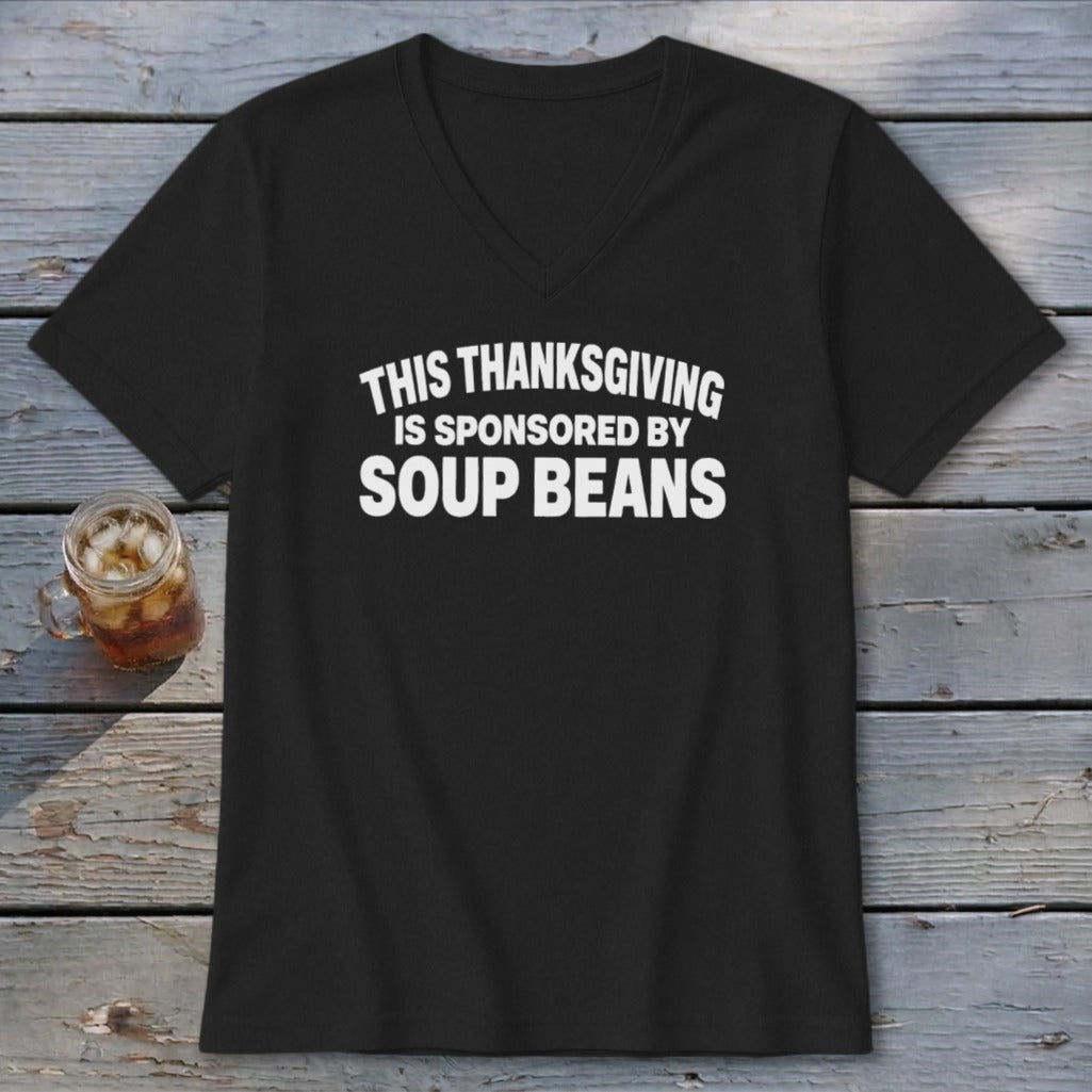 Black v-neck t-shirt featuring the white text "THIS THANKSGIVING IS SPONSORED BY SOUP BEANS" lying on a wooden surface.