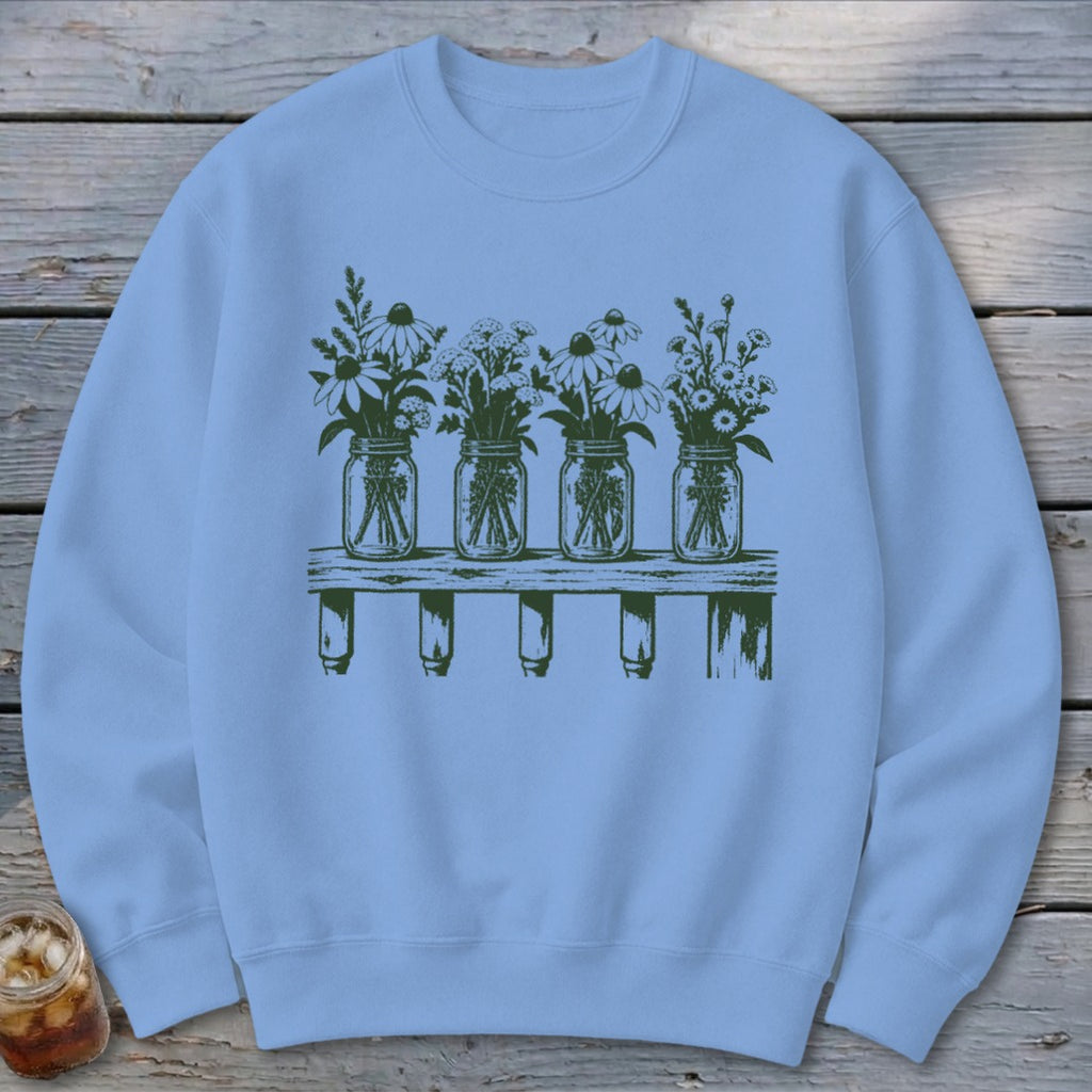 Light blue crewneck sweatshirt featuring a dark green graphic of four mason jars filled with wildflowers sitting on a wooden shelf.