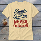 A light yellow t-shirt with blue and red text reading Sugar in my Sweet Tea NEVER IN MY CORNBREAD lies flat on a weathered wooden surface next to a glass of iced tea.