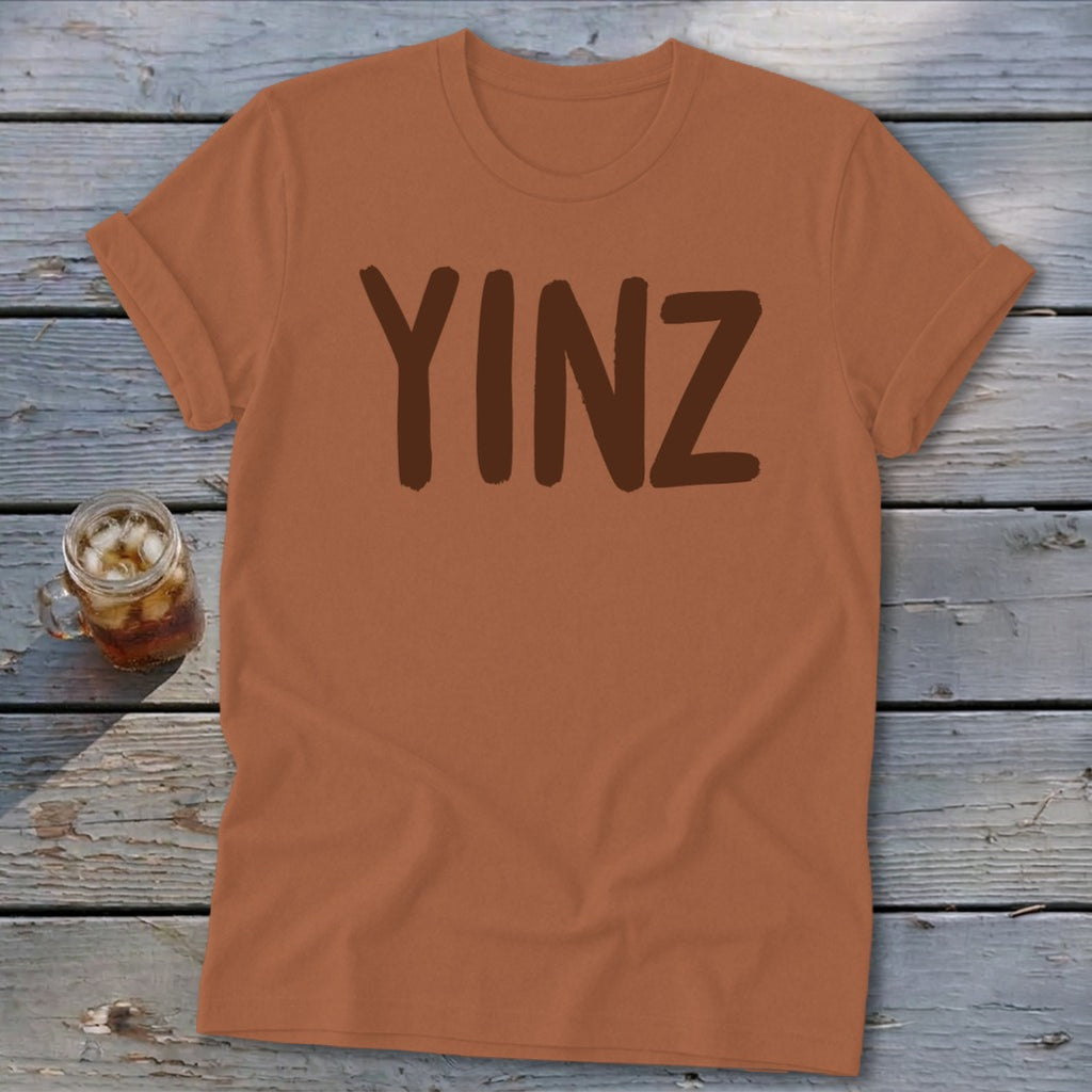 Brown t-shirt with "YINZ" printed in dark brown on a wooden background.