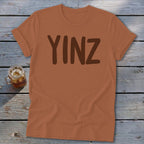 Brown t-shirt with "YINZ" printed in dark brown on a wooden background.