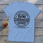 A light blue t-shirt printed with a circular badge featuring the text Certified Cornbread Judge and a cornbread illustration.