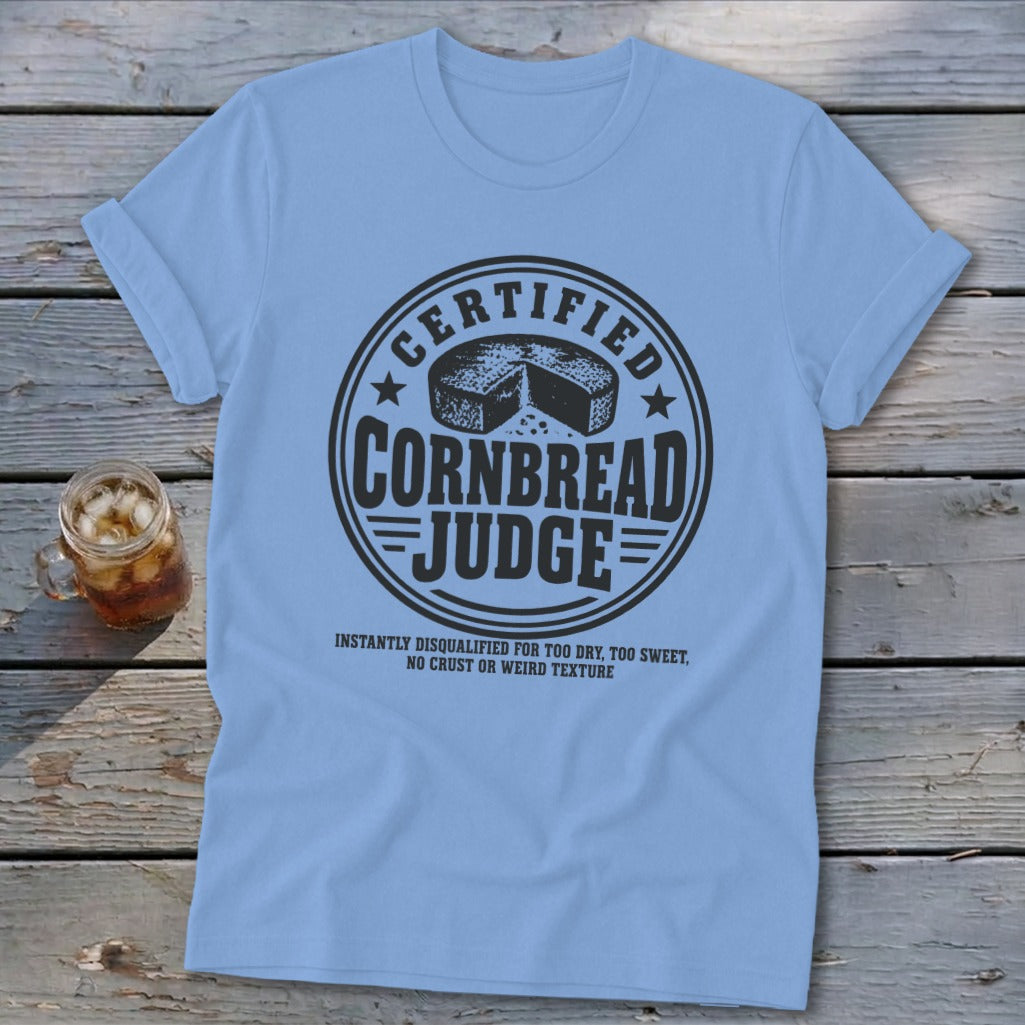 A light blue t-shirt printed with a circular badge featuring the text Certified Cornbread Judge and a cornbread illustration.