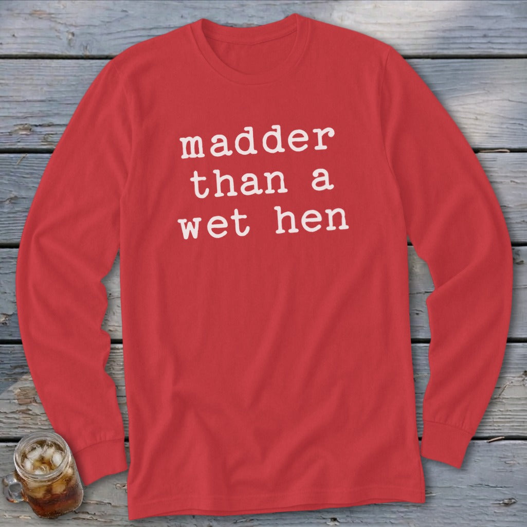 A red long-sleeve shirt with the white text "madder than a wet hen" on a wooden background.