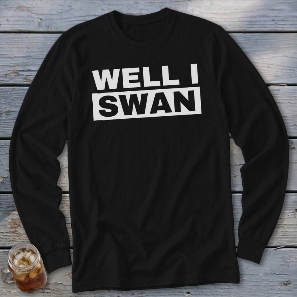 A black long-sleeve t-shirt with white text reading "WELL I SWAN" on a wooden surface.