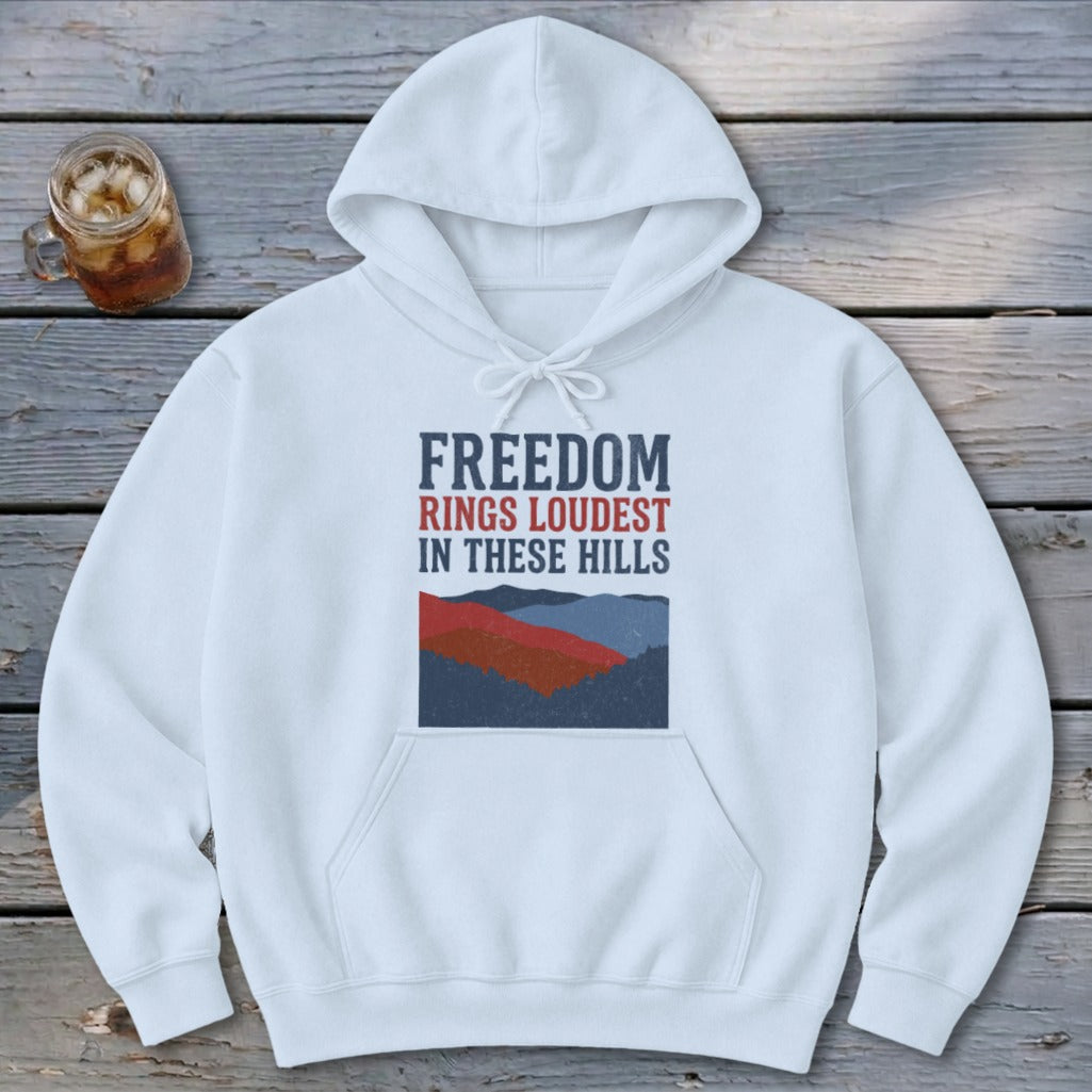 White hooded sweatshirt featuring blue and red text reading Freedom Rings Loudest in These Hills above a stylized mountain graphic.