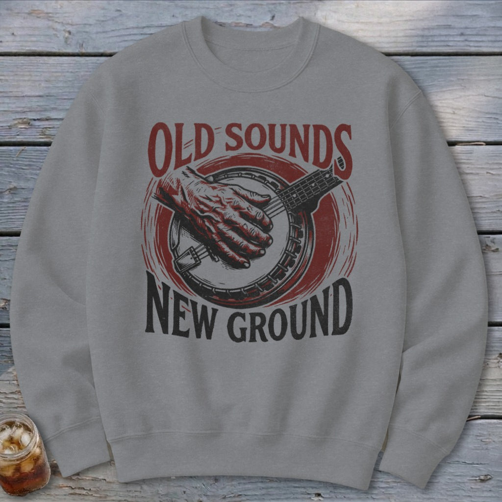 A gray crewneck sweatshirt printed with a red and black illustration of a hand playing a banjo and text reading Old Sounds New Ground.