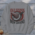 A gray crewneck sweatshirt printed with a red and black illustration of a hand playing a banjo and text reading Old Sounds New Ground.