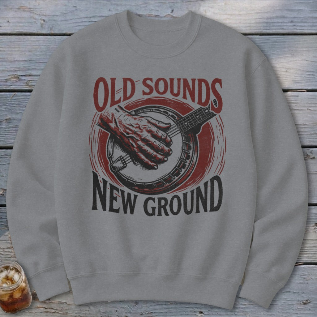 A gray crewneck sweatshirt printed with a red and black illustration of a hand playing a banjo and text reading Old Sounds New Ground.