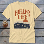 A cream-colored t-shirt with brown text reading "HOLLER LIFE" above a mountain and sun graphic.