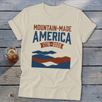 A cream-colored t-shirt with rolled sleeves lies on a rustic wooden table, featuring blue and red mountain graphics and text that reads "MOUNTAIN-MADE AMERICA 1776-2026."