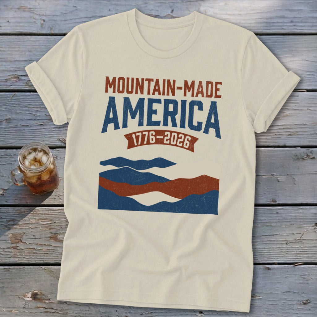 A cream-colored t-shirt with rolled sleeves lies on a rustic wooden table, featuring blue and red mountain graphics and text that reads "MOUNTAIN-MADE AMERICA 1776-2026."