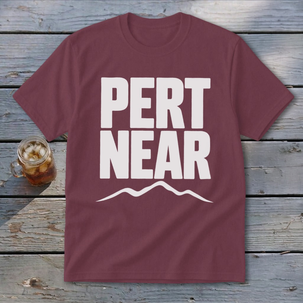 Maroon crewneck t-shirt featuring the white text "PERT NEAR" and a mountain silhouette, lying on a wooden surface.