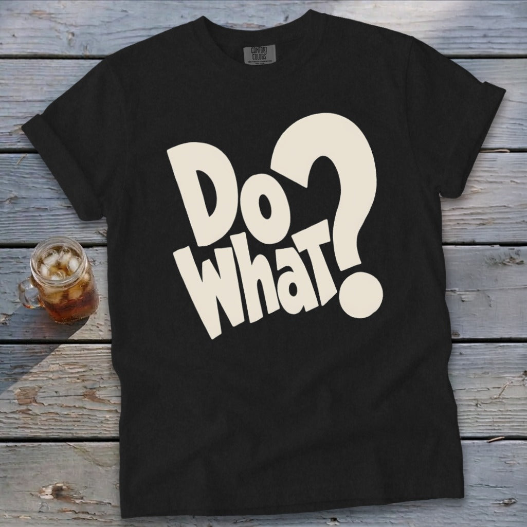 A black t-shirt with rolled sleeves features beige text that reads "DO WHAT?" displayed on a rustic wooden table next to a glass of iced tea.