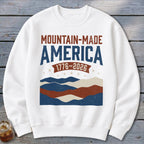 A white crewneck sweatshirt featuring a graphic with the text Mountain-Made America 1776-2026 above abstract mountains.