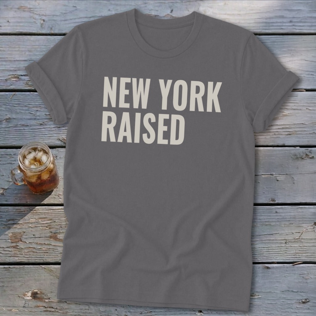 Gray t-shirt with "NEW YORK RAISED" text in white, next to a glass of iced drink.