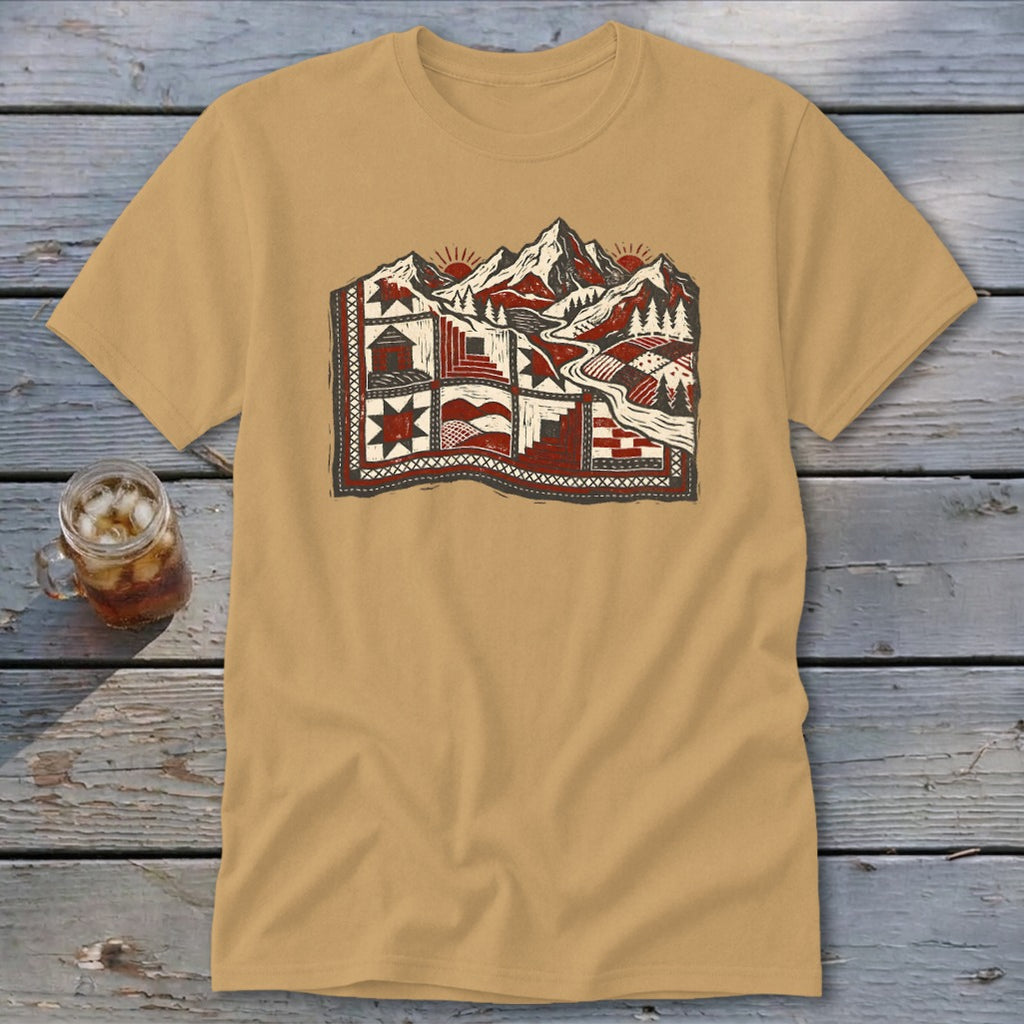 Tan t-shirt featuring a stylized graphic of snowy mountains and a quilt pattern on a wooden surface.