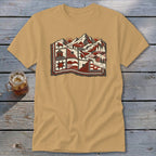 Tan t-shirt featuring a stylized graphic of snowy mountains and a quilt pattern on a wooden surface.