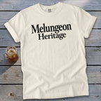 Off-white t-shirt with black text reading "Melungeon Heritage" laid flat on a rustic wooden surface next to a glass of iced tea.