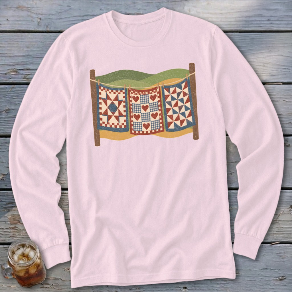 Light pink long-sleeve t-shirt with a vintage graphic of three quilts hanging on a clothesline against rolling hills.